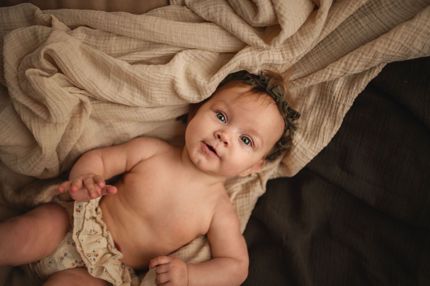 Baby in cream colored clothing reclining on textured neutral backdrop.