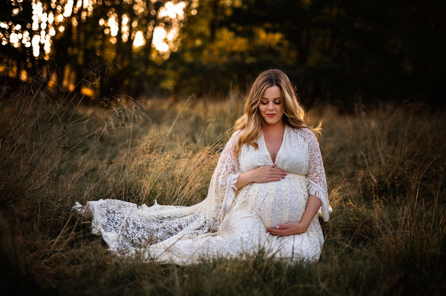 Elegant maternity photo in white lace dress amid golden evening light.