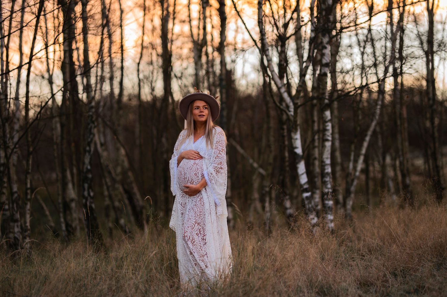 Figure in white lace dress stands in tall grass during sunset.