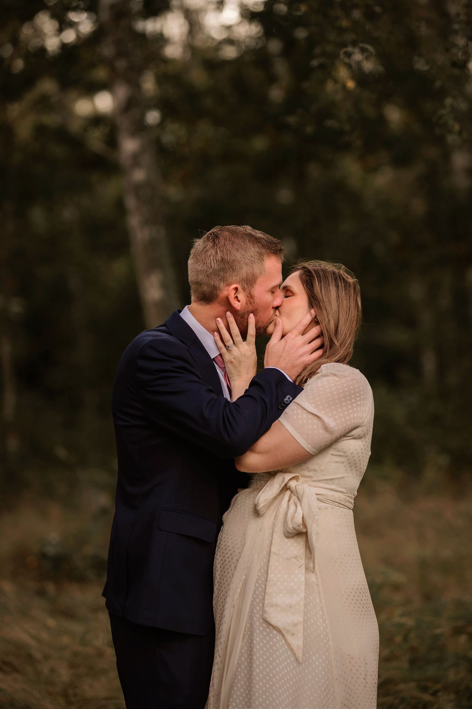 Couple sharing a romantic kiss in a forest, man in navy suit and woman in cream dress.
