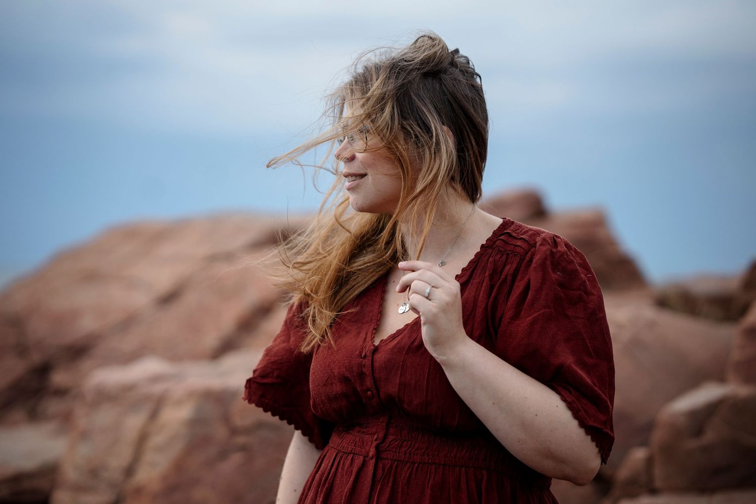 Woman in red dress with windswept hair poses among red rocks under a cloudy sky.