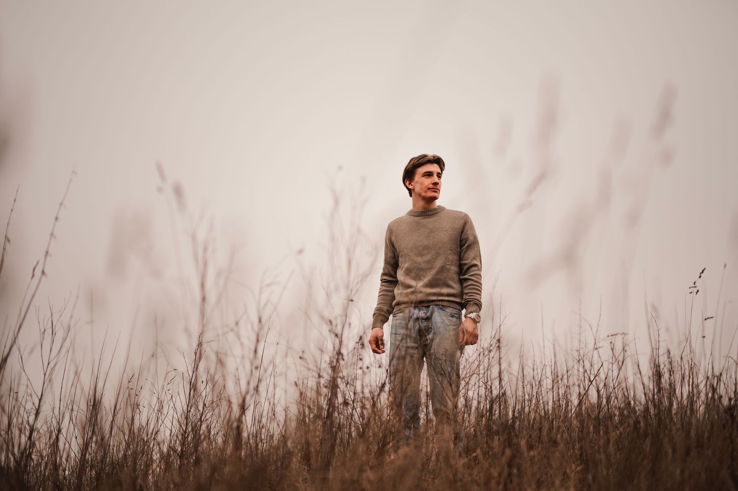 Young man standing alone in a misty field of tall dry grass wearing a beige sweater.