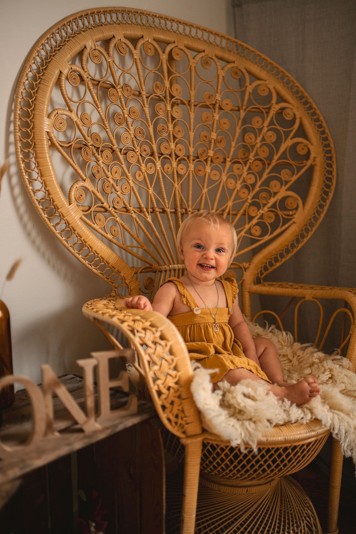 Baby smiles while seated in ornate rattan peacock chair with rustic ONE decoration.