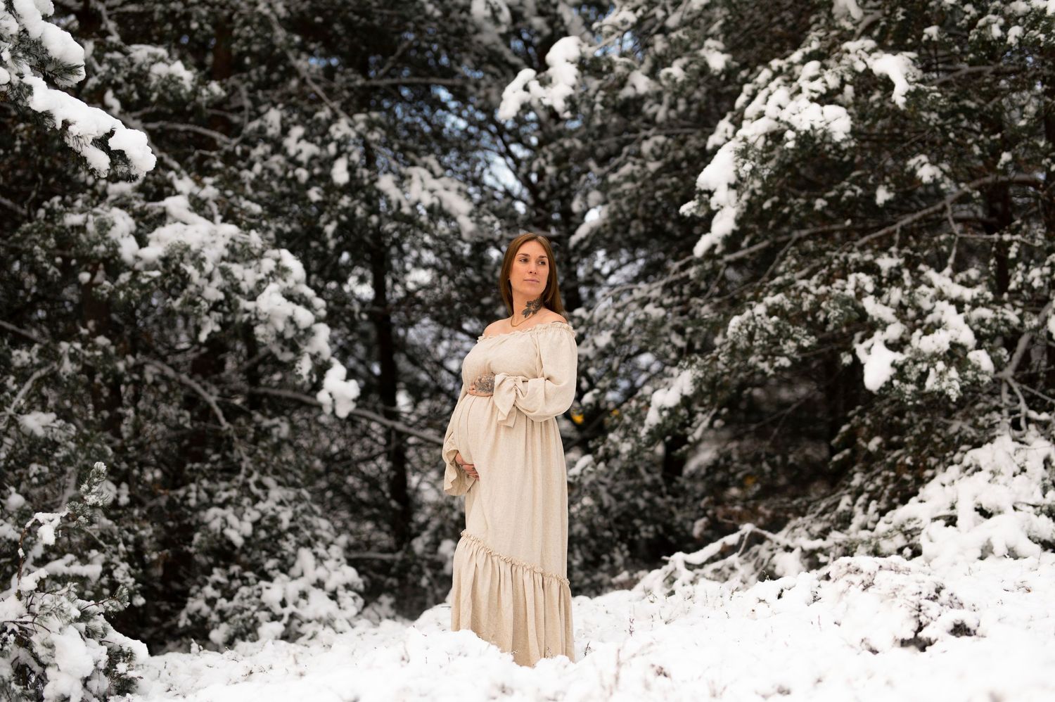 Elegant maternity portrait in a snow-covered forest with evergreen trees.