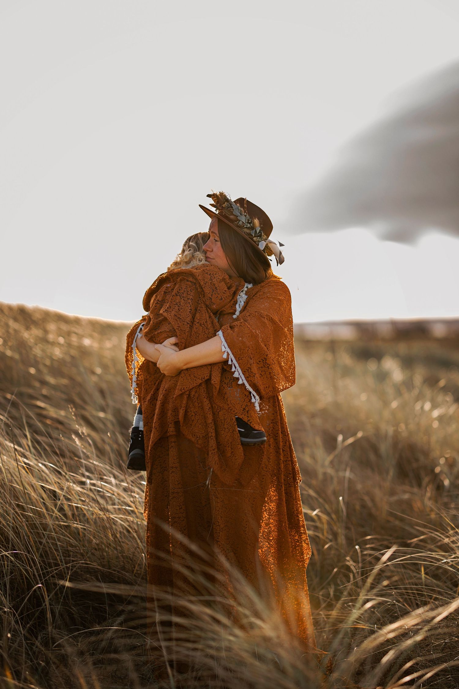 Rustic maternity portrait in brown dress and hat amid golden prairie grasses at sunset.