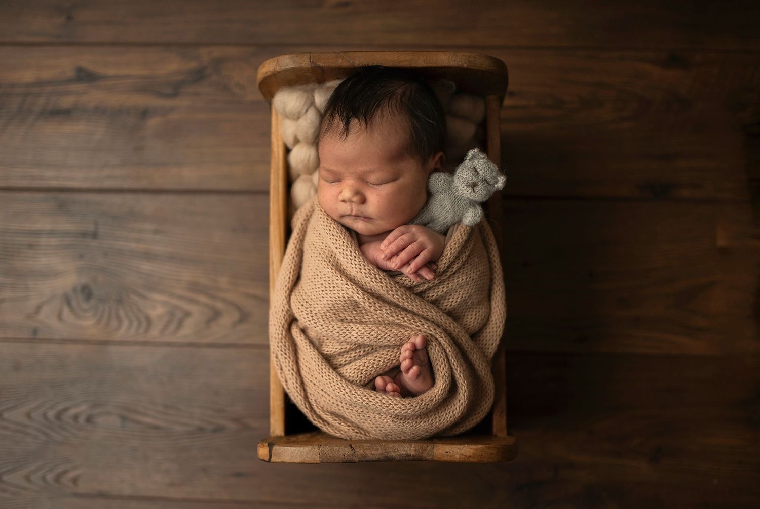 Newborn sleeps peacefully in beige wrap on wooden bed with small stuffed animal.