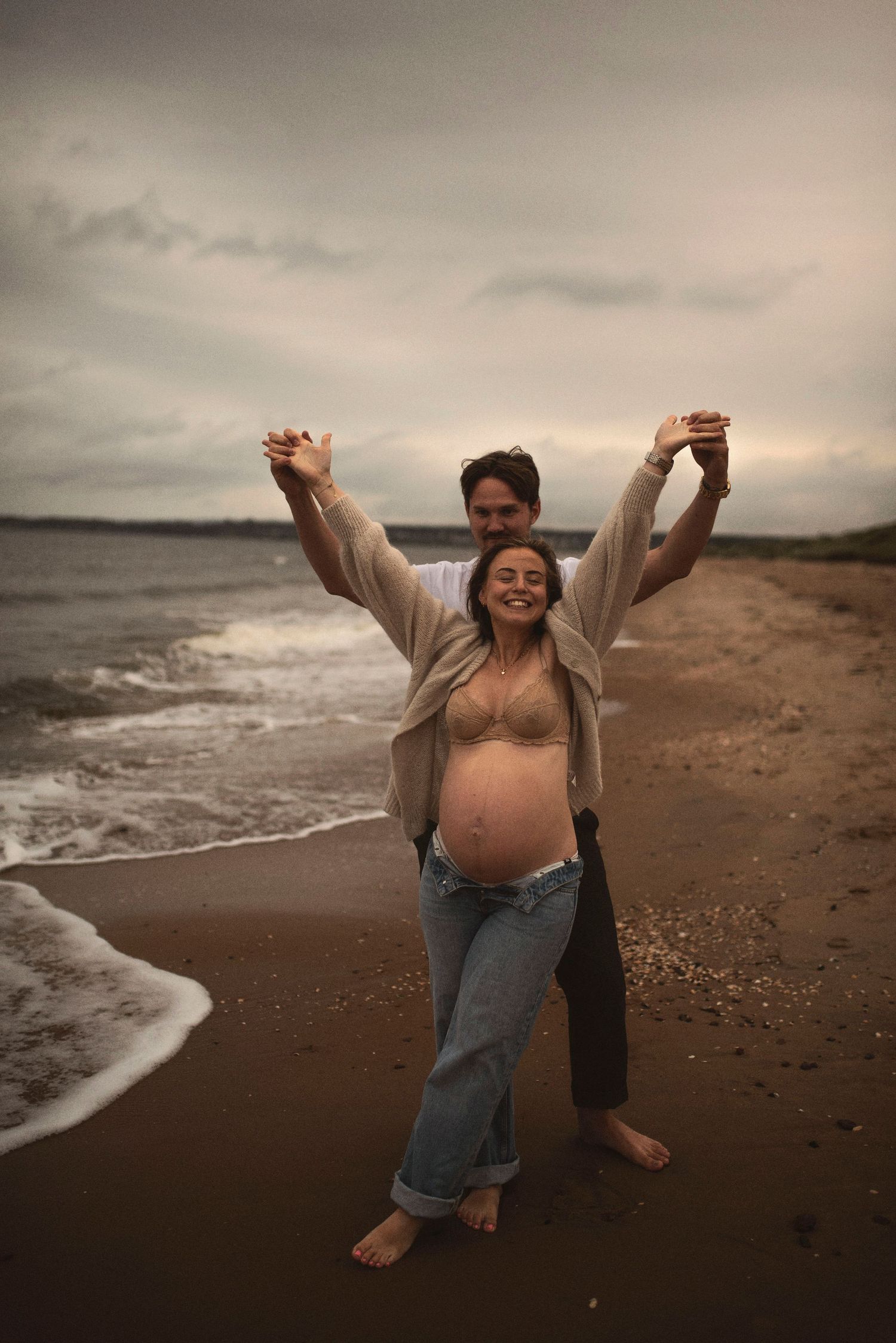 Couple shares a playful moment on the beach during sunset with arms raised.
