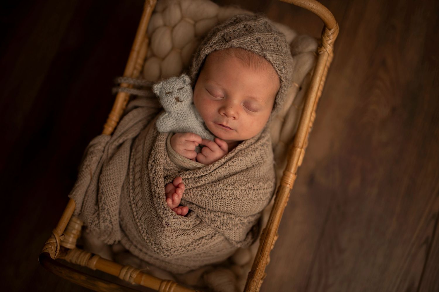 Sleeping newborn wrapped in beige knit blanket with small toy in wicker basket.