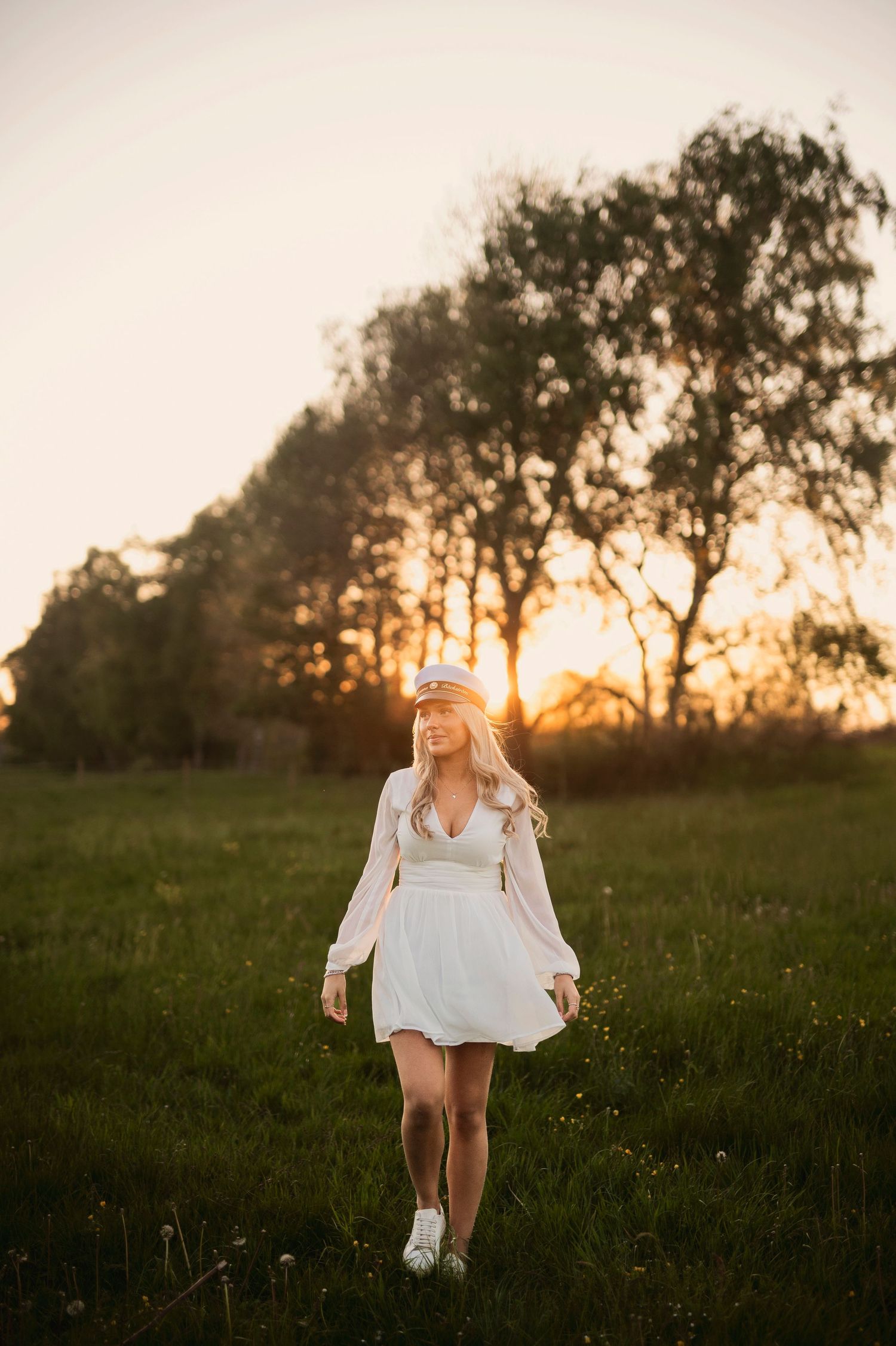 Blonde woman in white dress and captain hat walking through a sunlit green meadow.