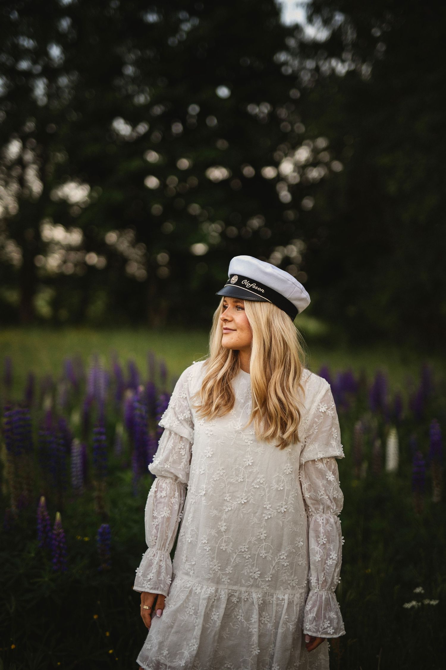 Blonde woman in white lace dress and captain hat standing in a field of purple lupines.