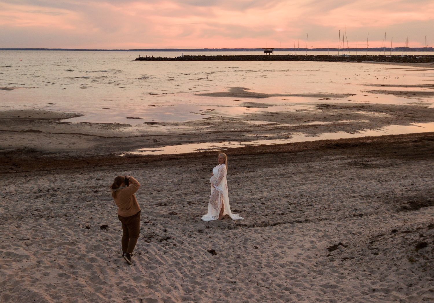 Photographer capturing a woman in white dress on sandy beach during pink sunset in Skåne, Sweden.
