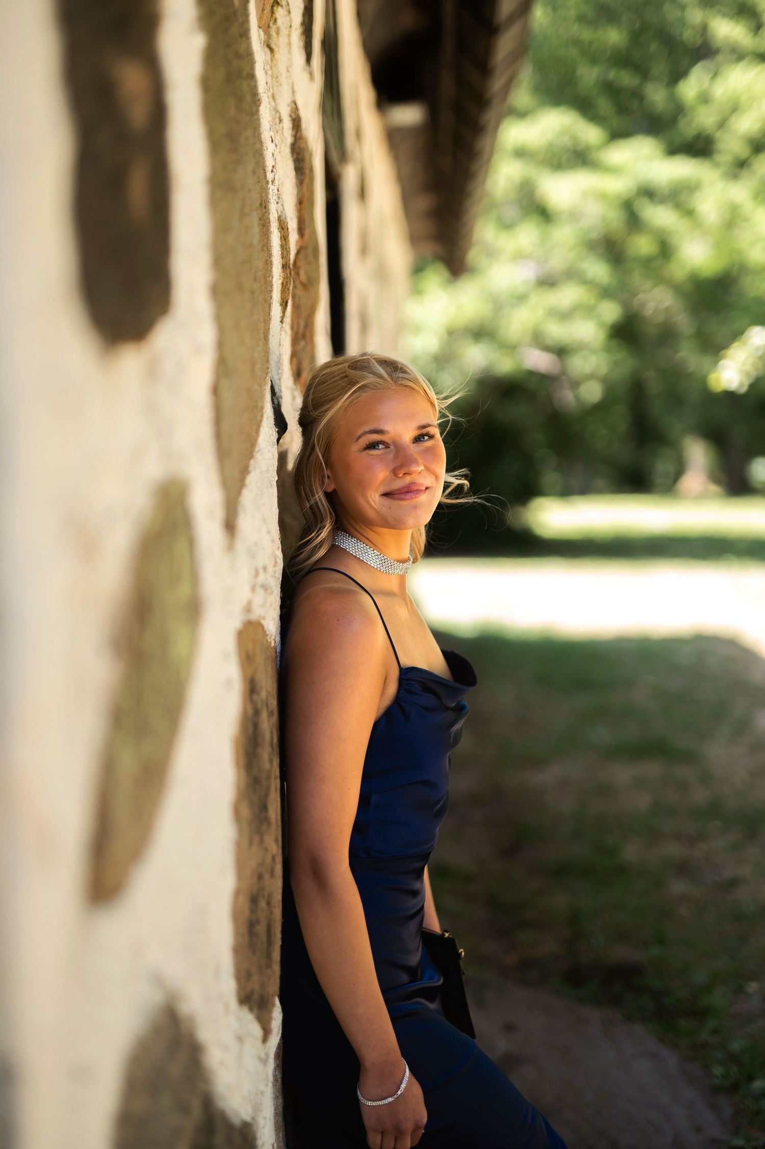 A smiling blonde woman in a navy blue dress leans against a stone wall in a sunlit outdoor setting.