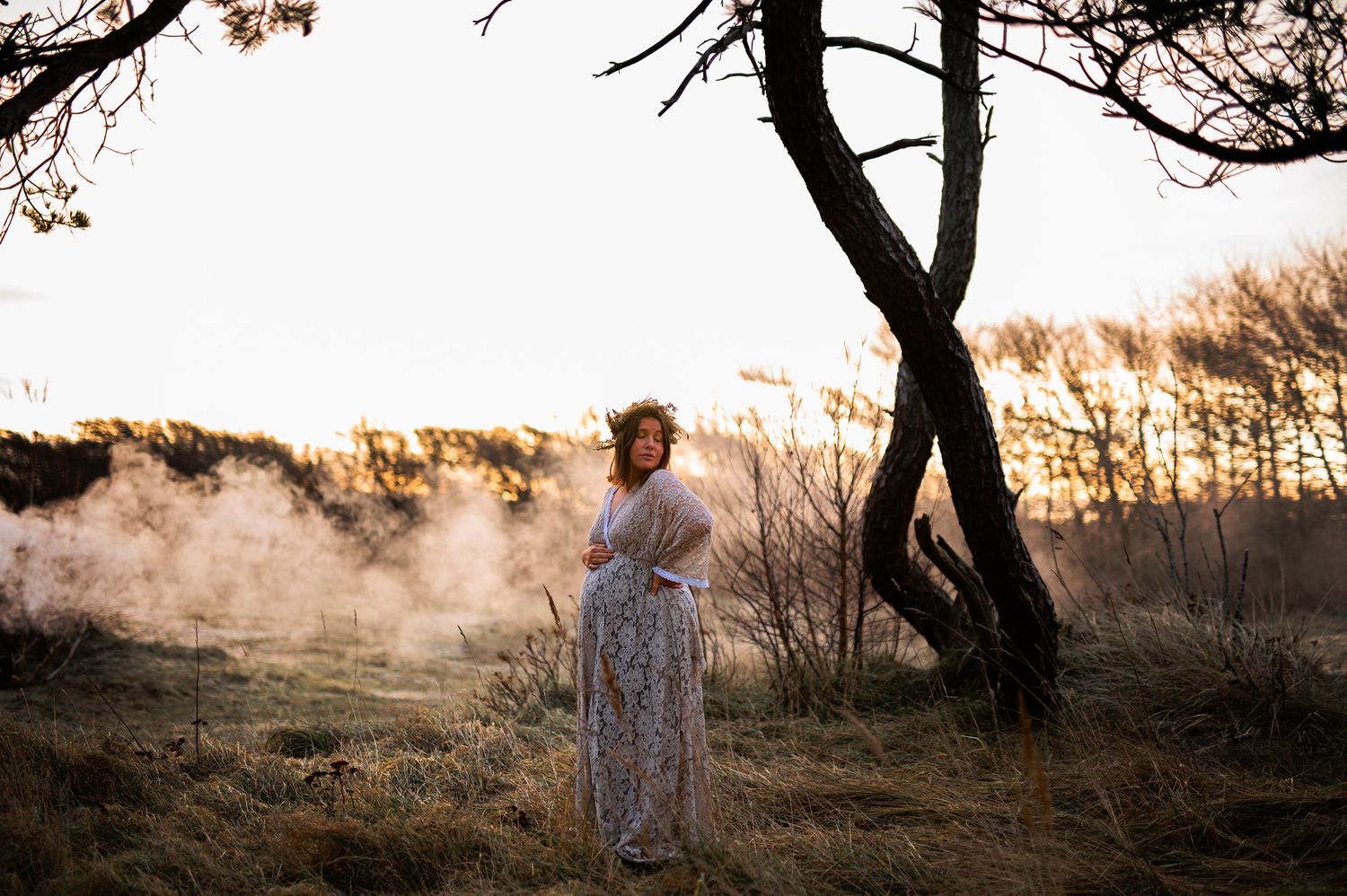 Ethereal maternity photograph with morning mist and twisted trees creating moody atmosphere.