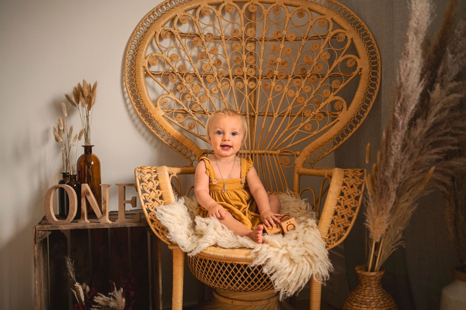 Baby in mustard dress sitting on peacock chair with sheepskin and rustic decor.