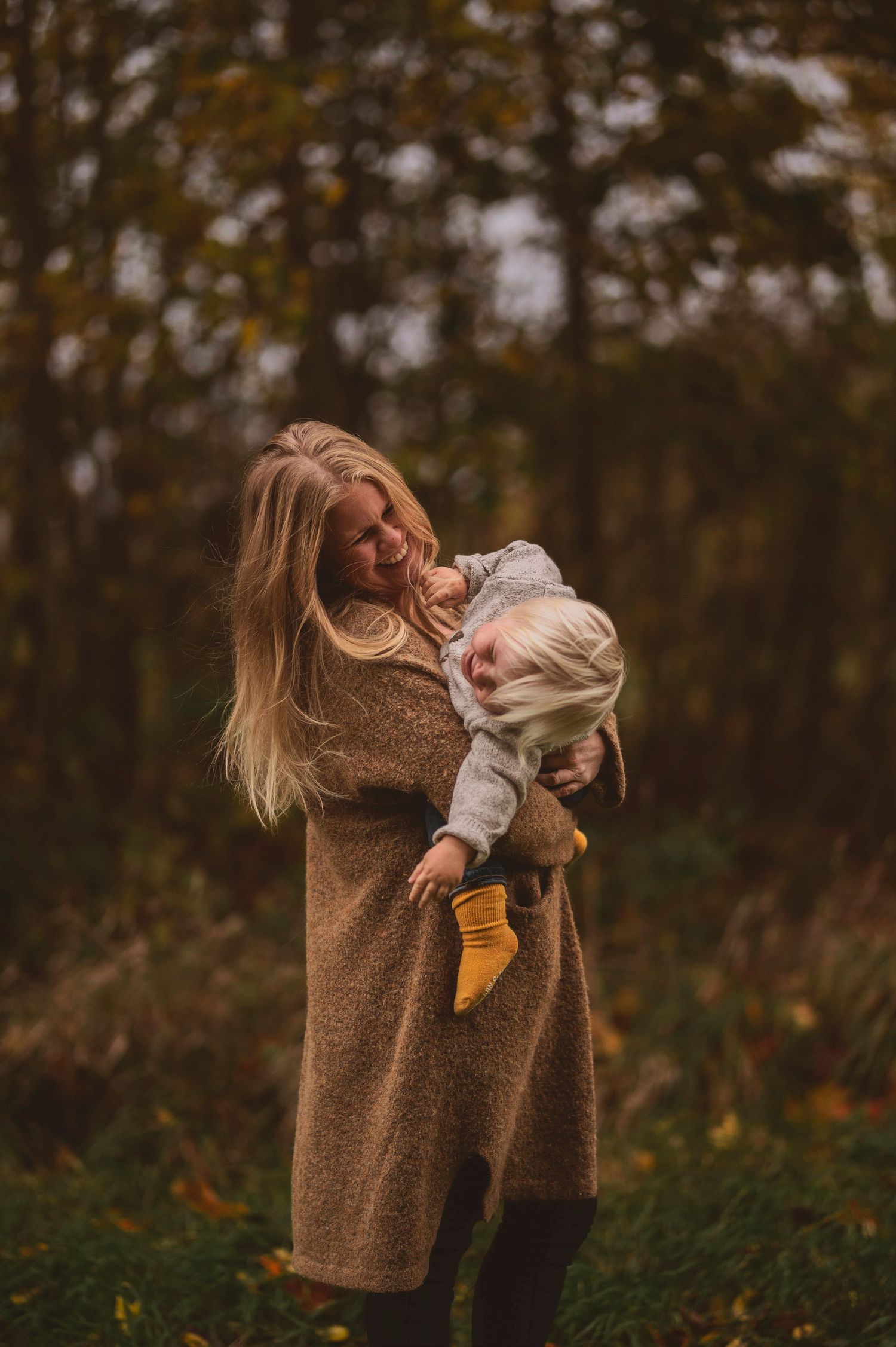 Adult in cozy brown sweater holds child wearing yellow socks in autumn outdoor setting.