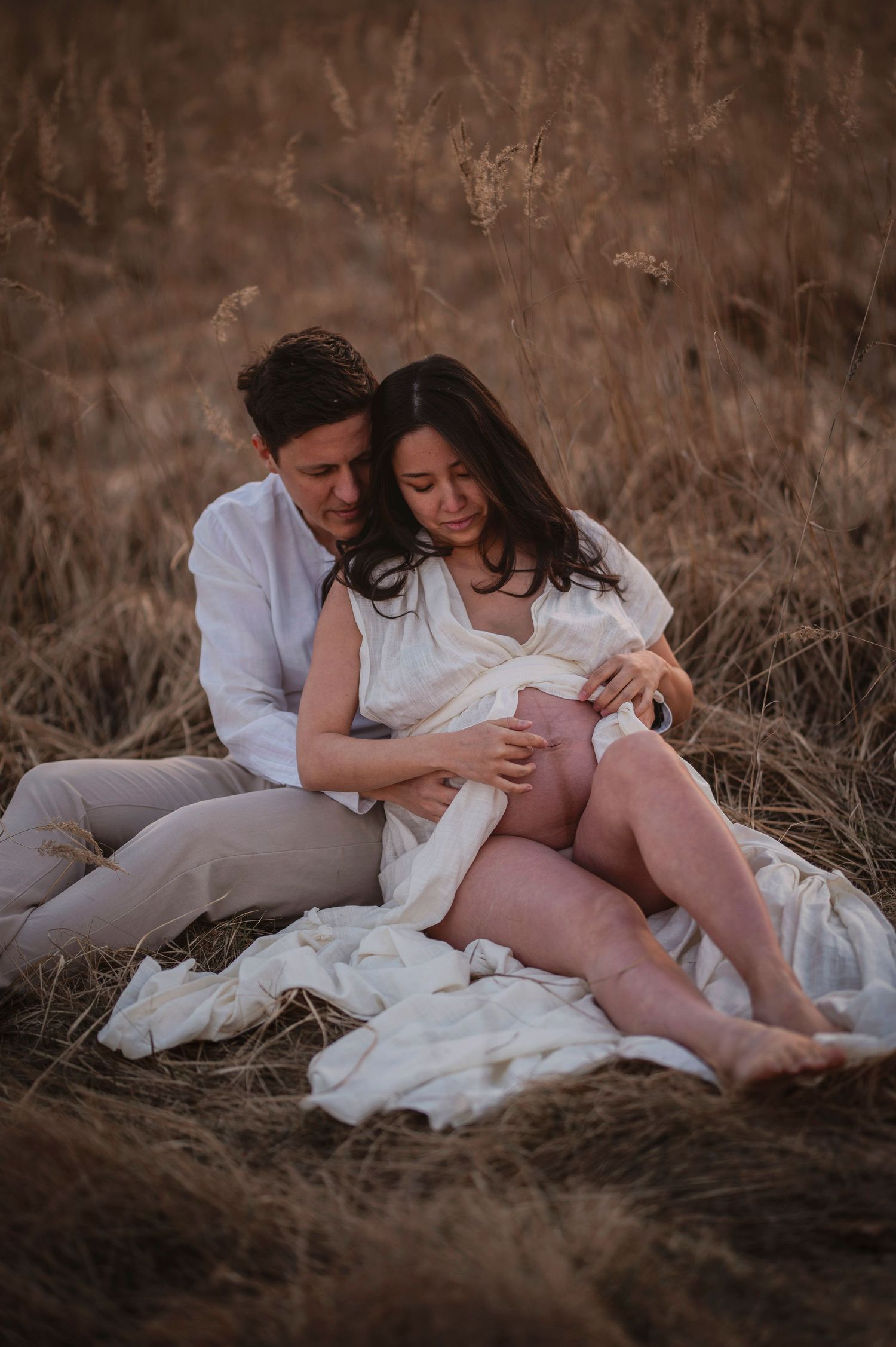 A romantic sepia-toned portrait of a couple in white attire sharing a tender moment while sitting in tall grass.