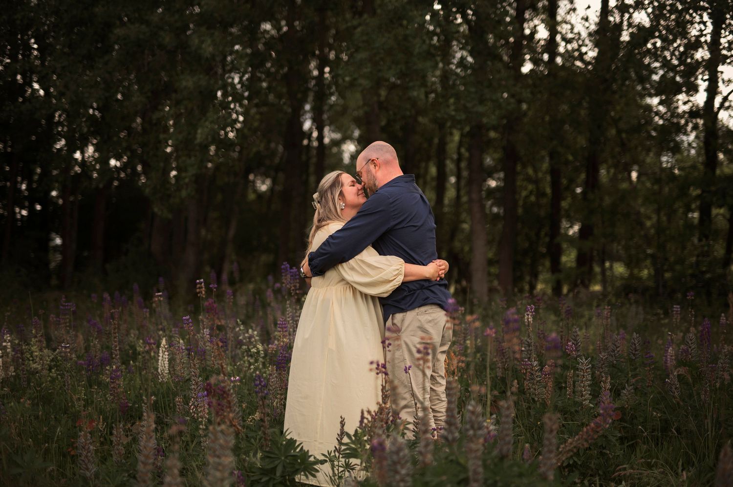 Couple embracing in cream colored outfits among purple wildflowers at sunset.