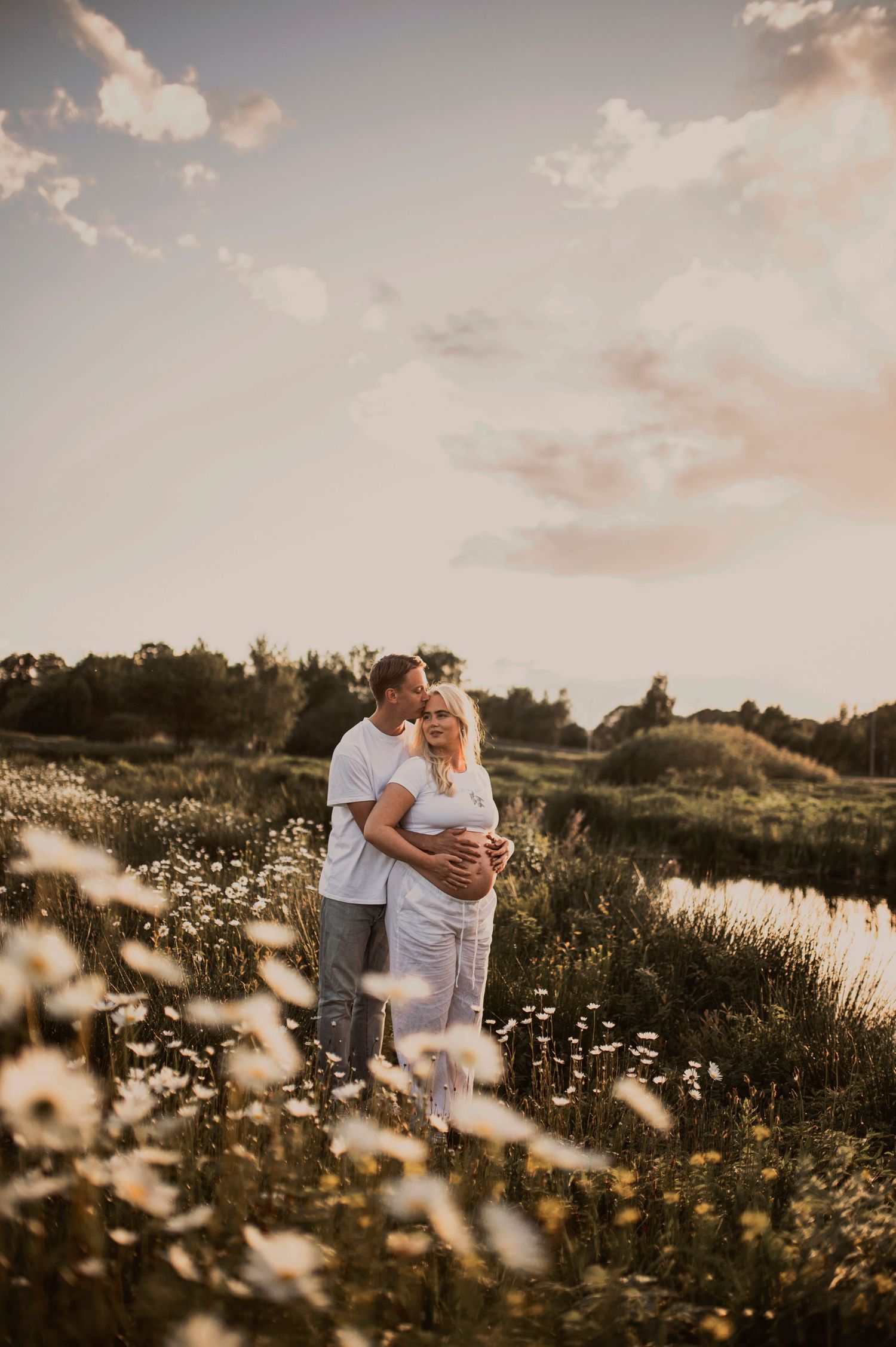 Couple embraces in a meadow of white wildflowers as the sun sets behind a tranquil pond.