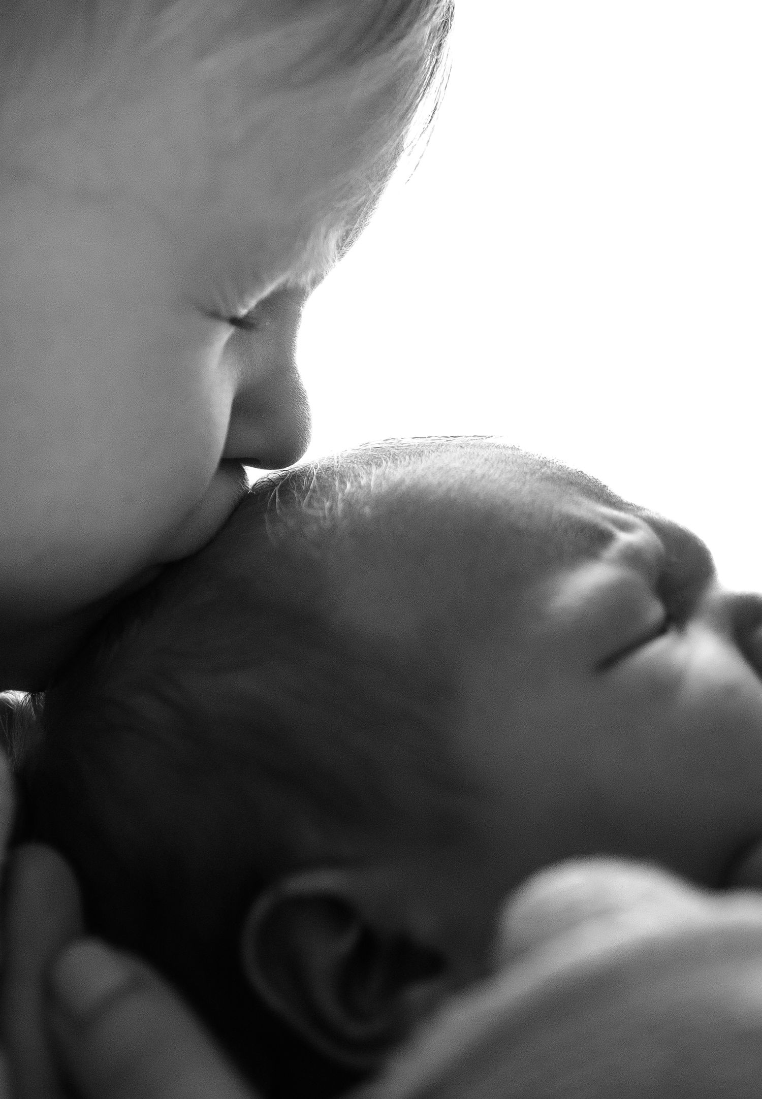 Black and white close-up of a young child gently kissing a sleeping newborn baby on the forehead.