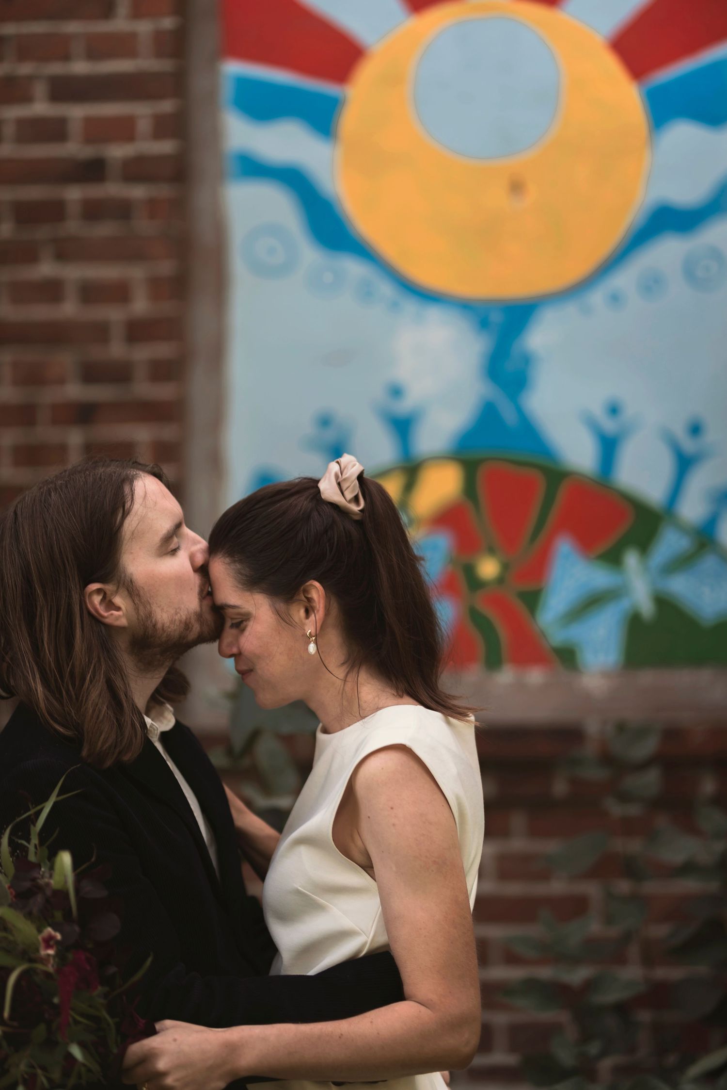 A groom tenderly kisses his bride on the forehead in front of a colorful sun mural at their wedding.