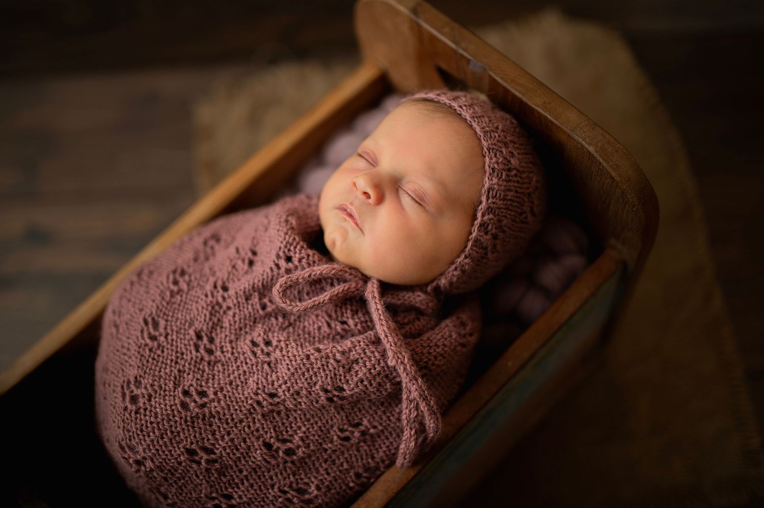 Peaceful sleeping newborn swaddled in mauve knit wrap in wooden basket.
