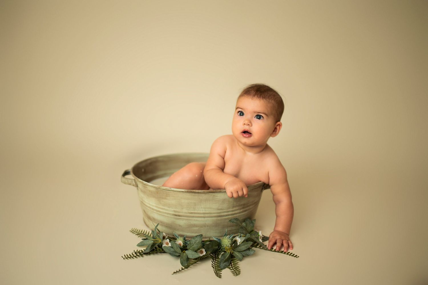 Baby sitting upright in metal basin surrounded by green foliage.