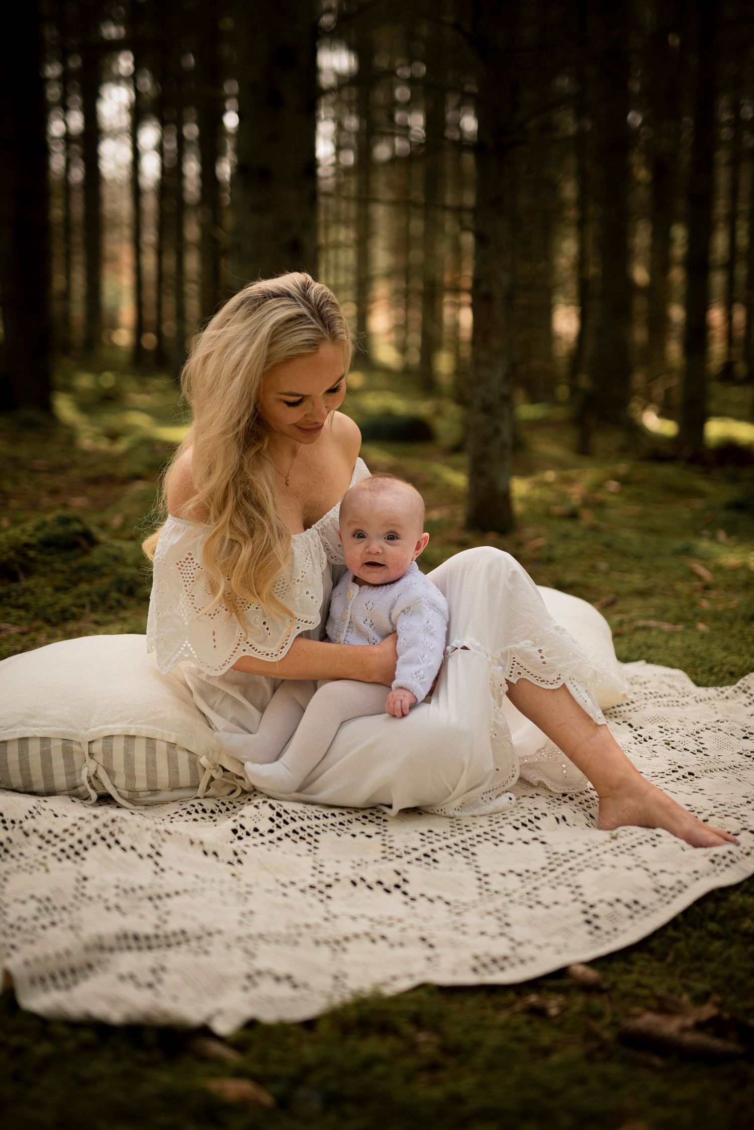 Mother and baby in white dress sitting on lace blanket in forest setting.