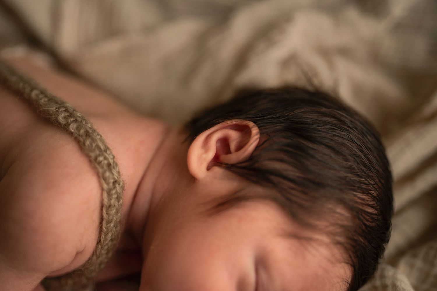 Sleeping newborn with dark hair rests on beige textured fabric in artistic lighting.