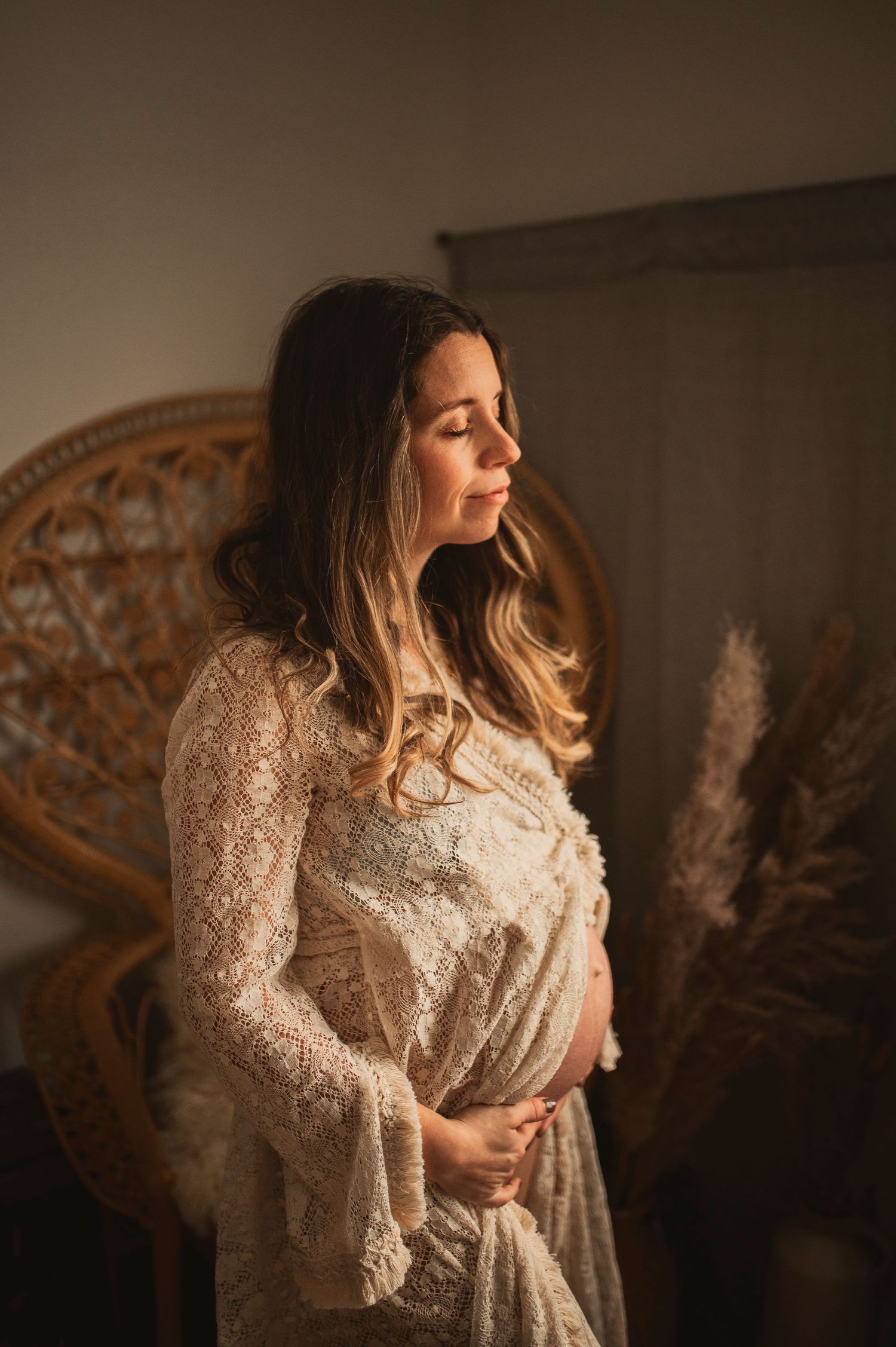 Woman in lace dress stands beside ornate vintage frame in warm lighting.