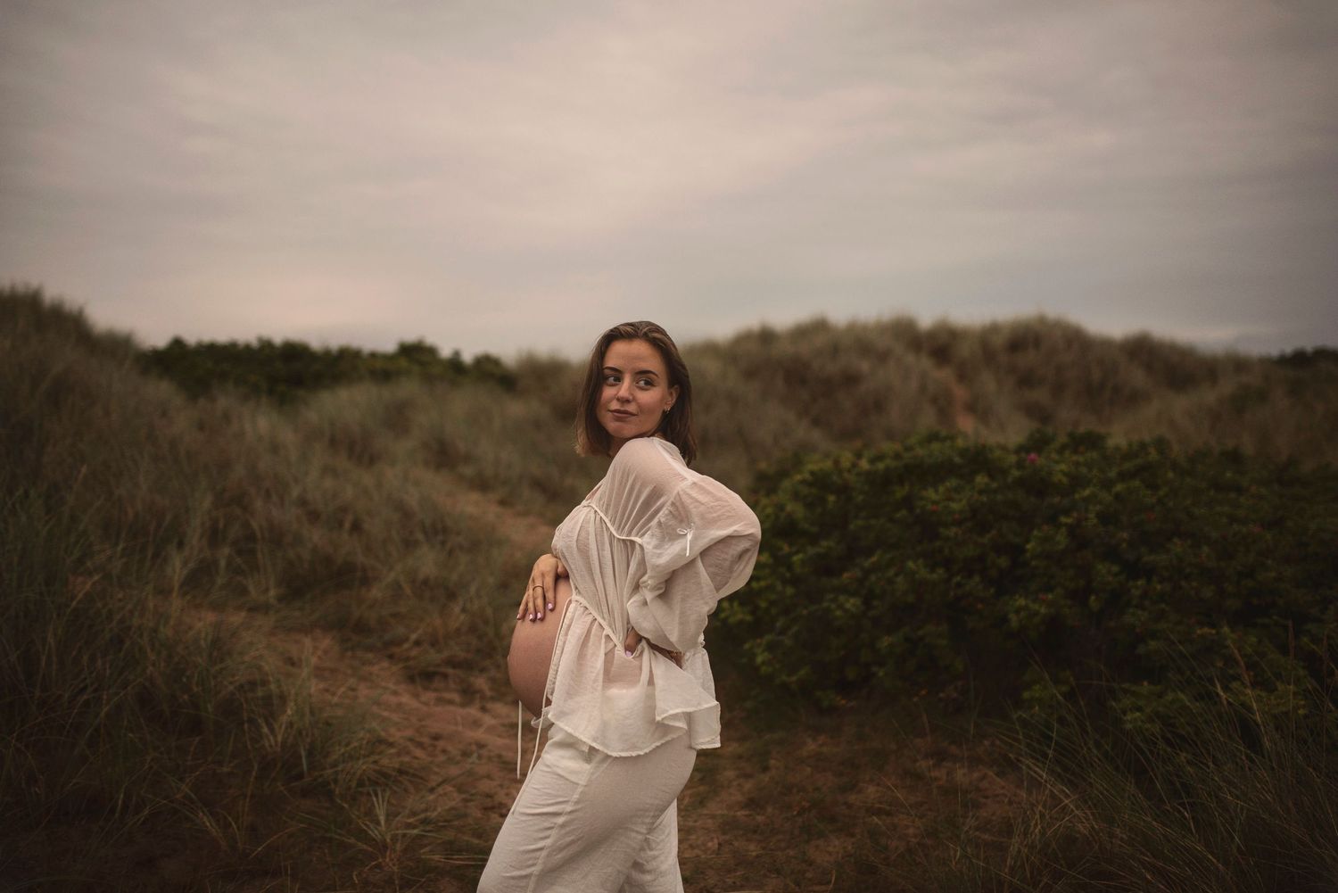 A woman in white clothing poses against a scenic coastal backdrop during sunset.