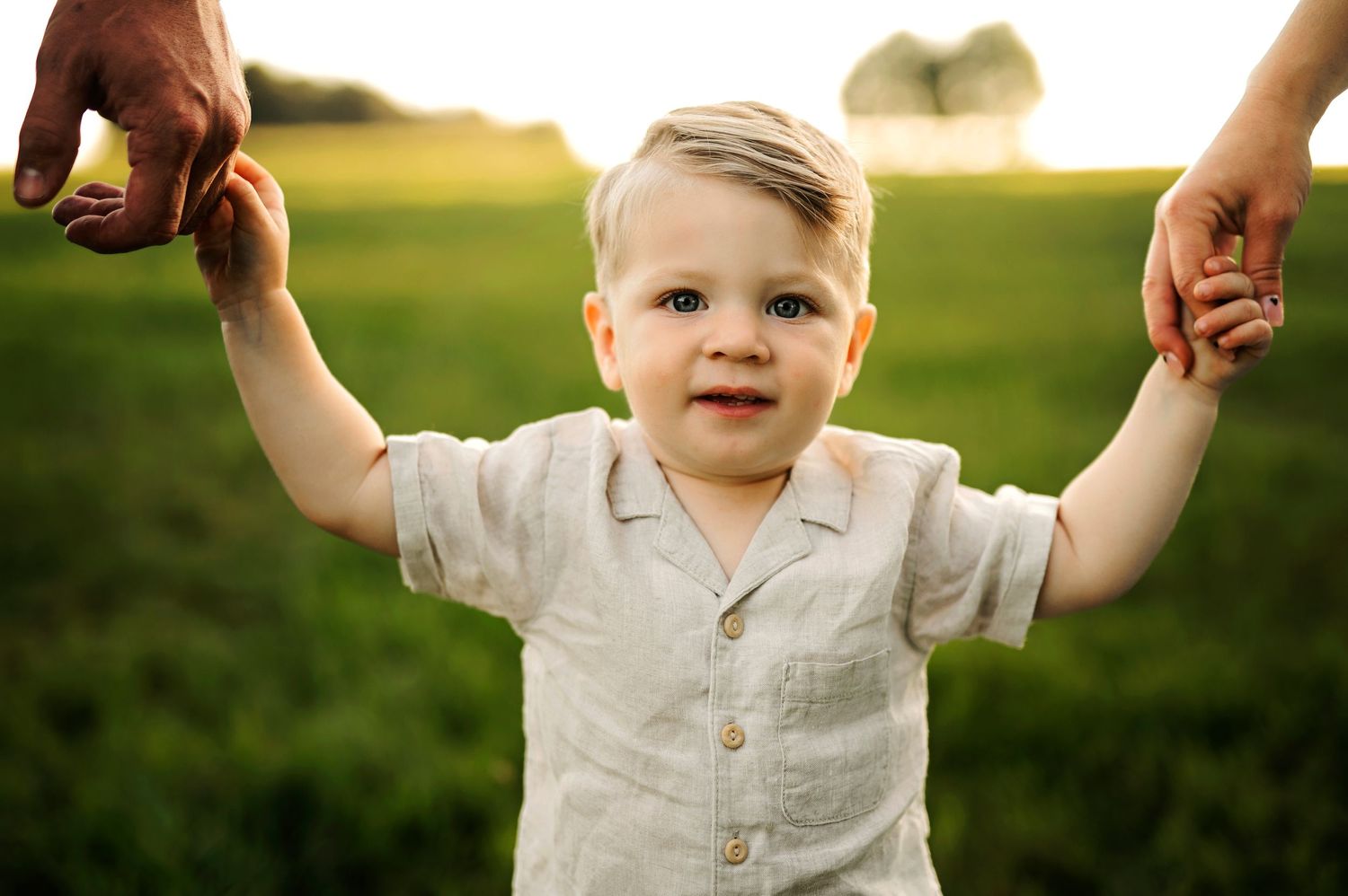 Toddler in white shirt smiling while holding hands with parents in a sunlit field.