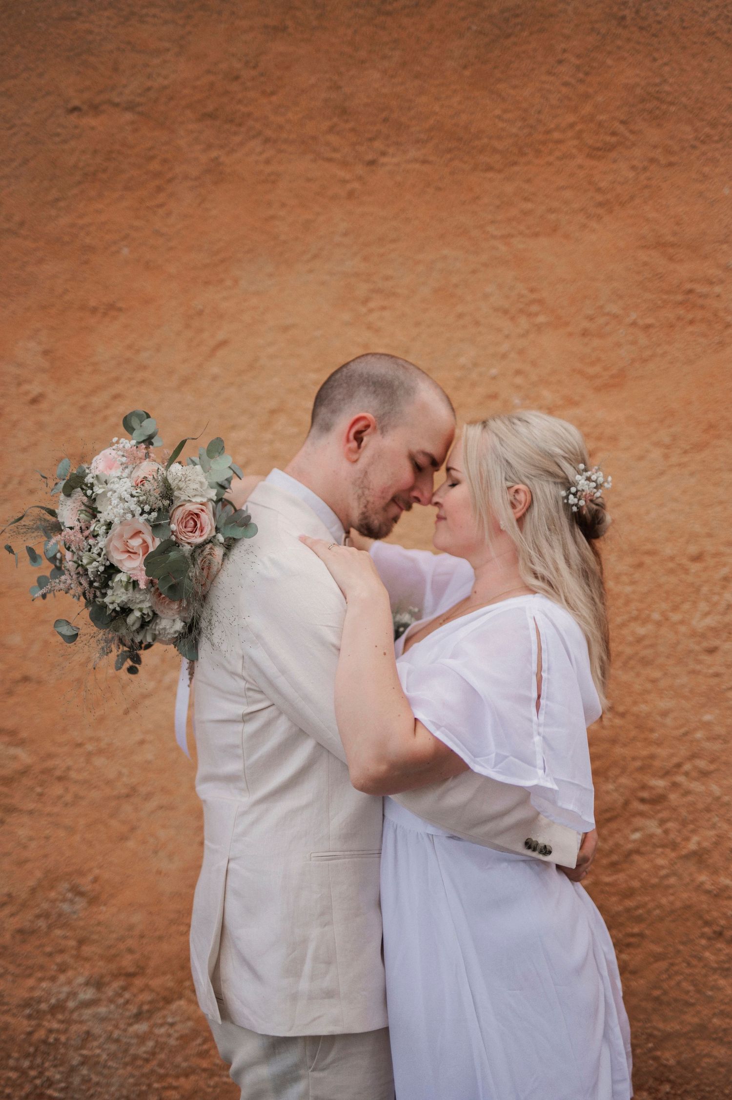 Wedding couple embracing against terracotta wall with pink and white floral bouquet.