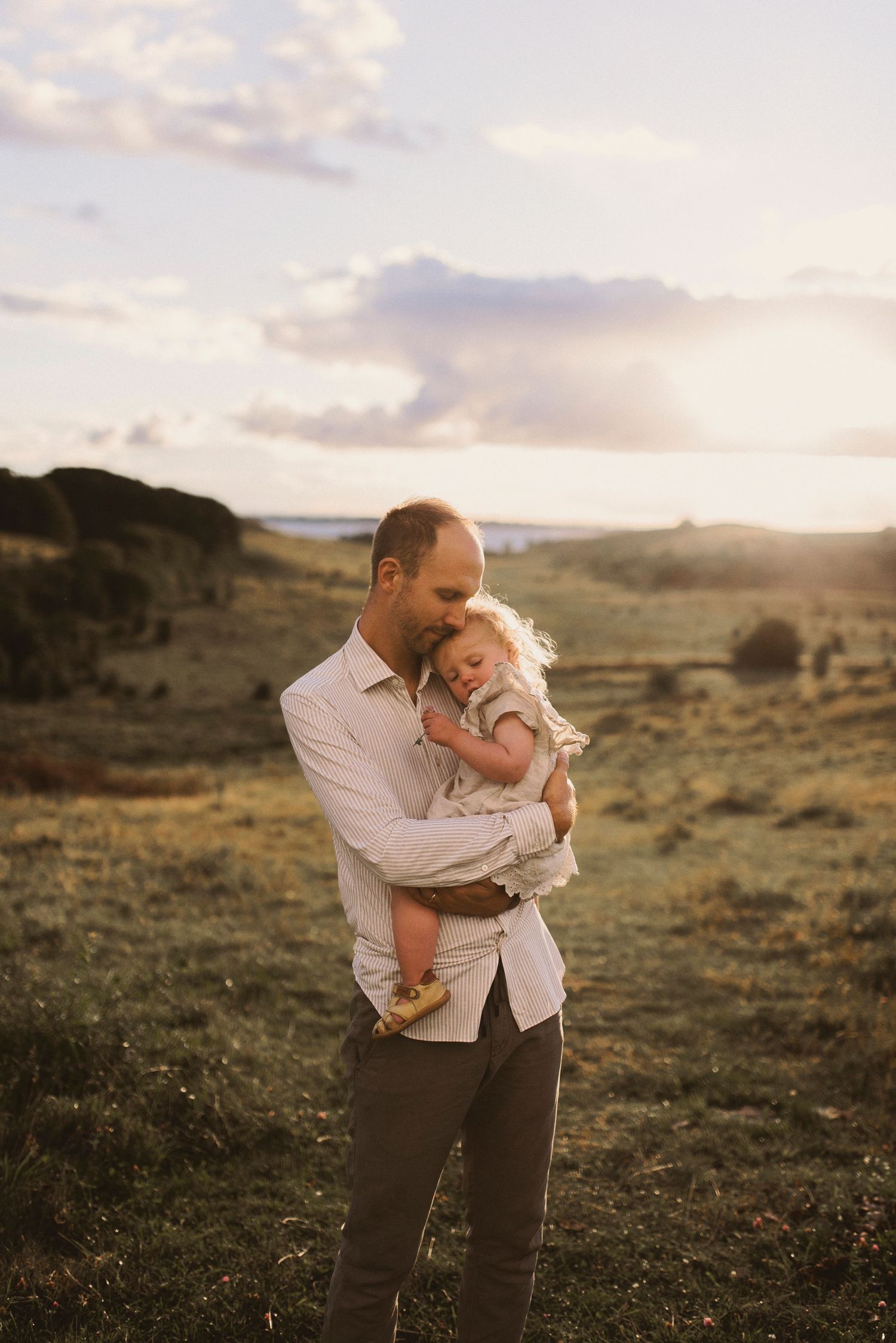 Parent holding infant against sunset landscape with rolling hills.