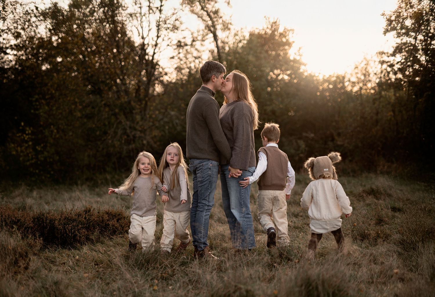 A family with four young children shares a golden hour moment in an autumn field, parents kissing while kids play around them.