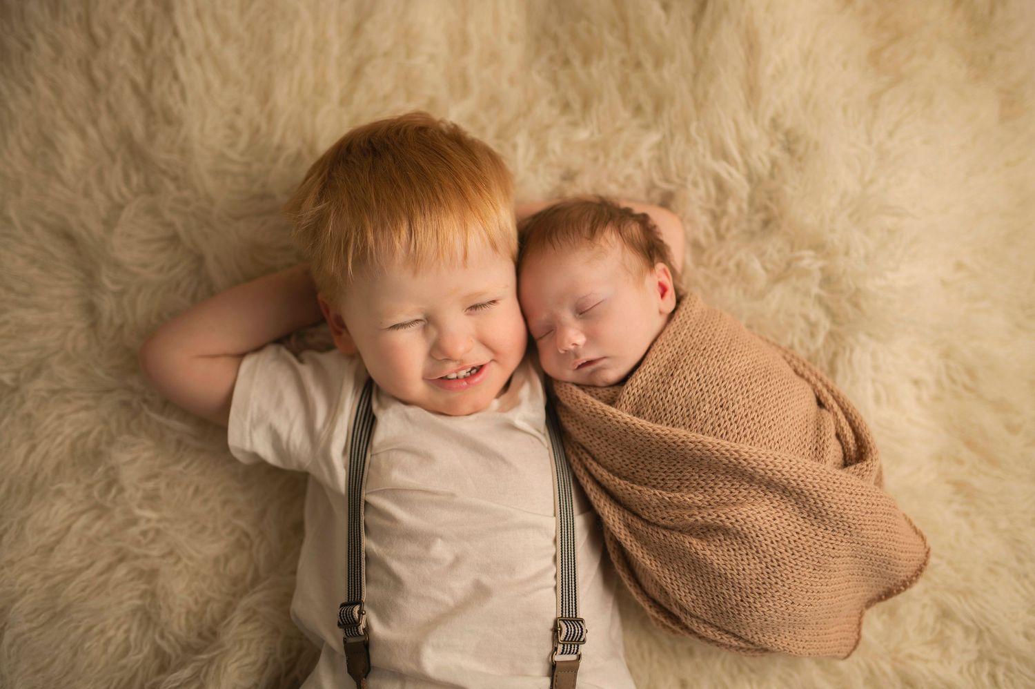 Two young children lying on a fluffy cream carpet, one wearing suspenders and one wrapped in a brown blanket.