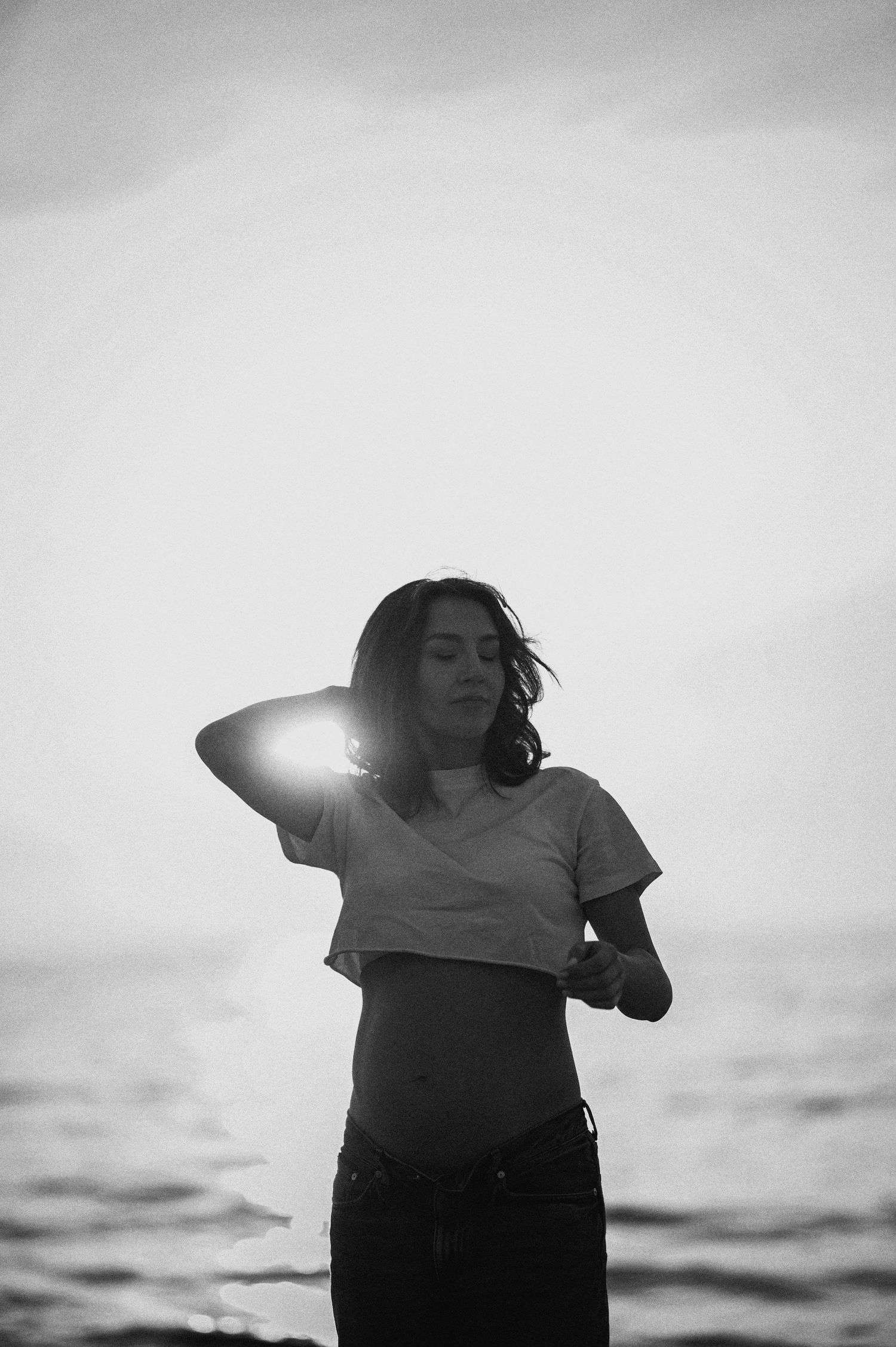 Black and white photo of a figure standing against sunlit ocean background in casual attire.