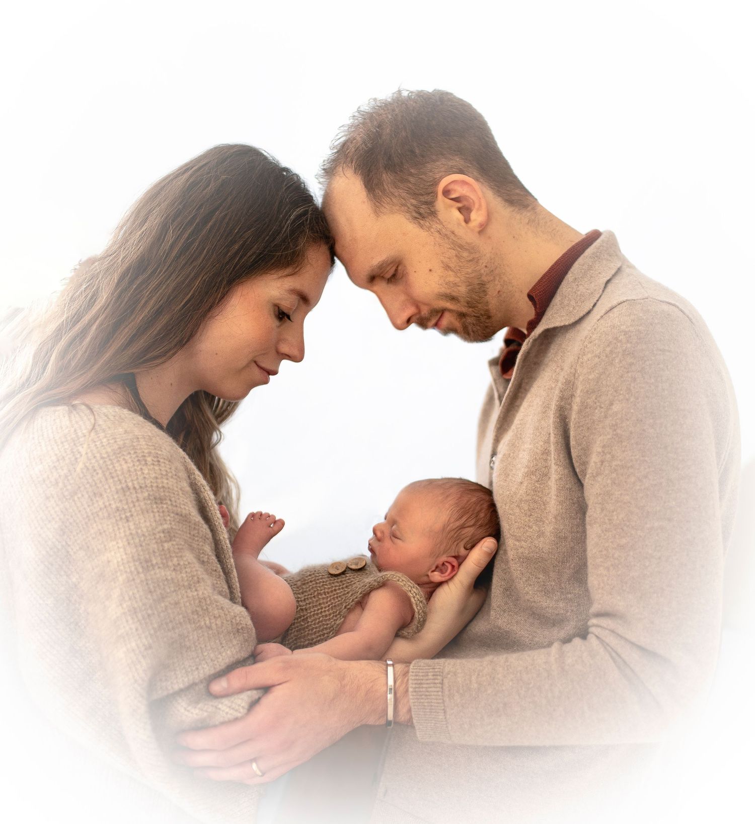 Intimate family moment with new parents holding their swaddled newborn against white background.