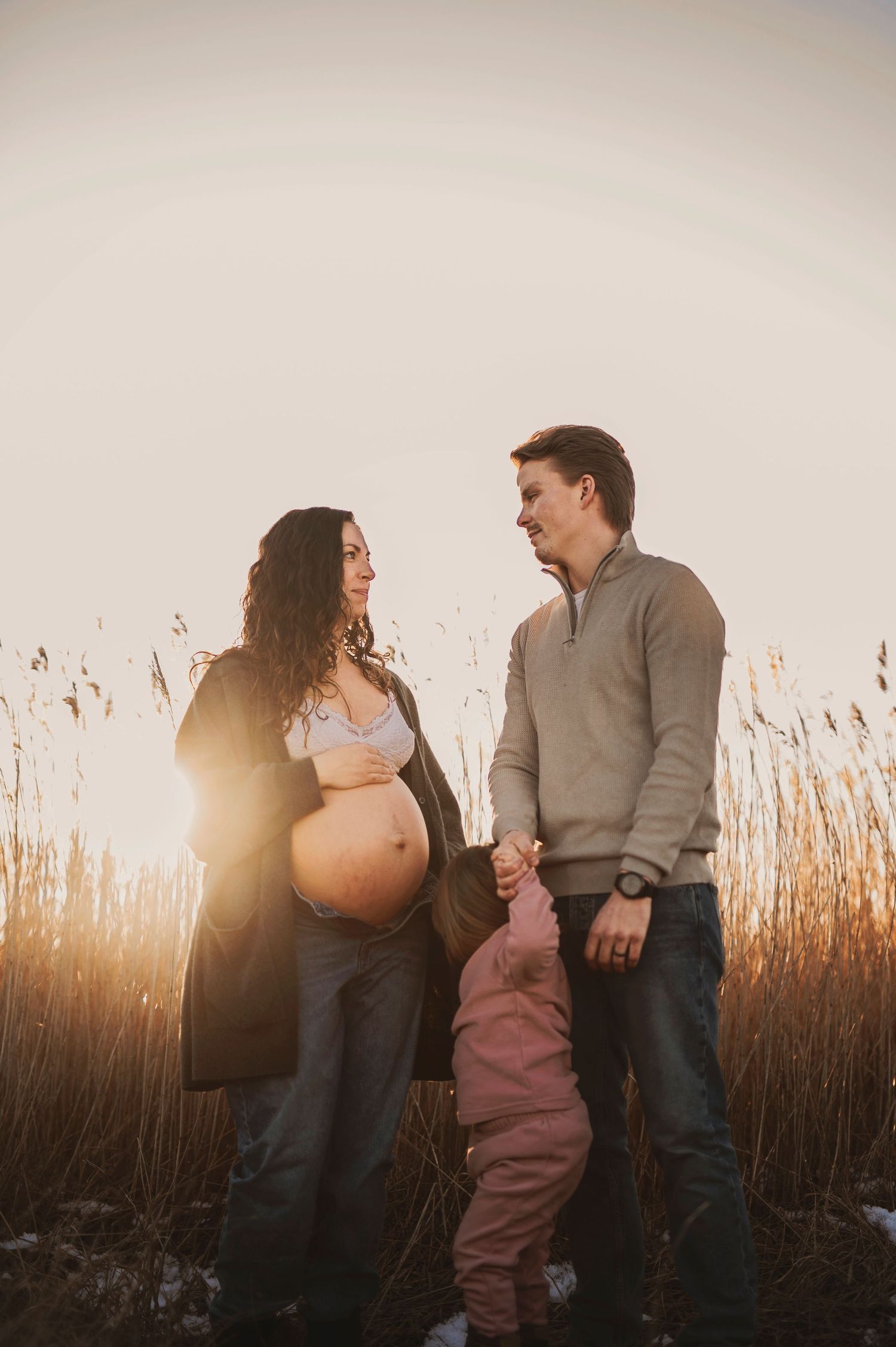 Maternity portrait of couple with child against sun-lit reeds in winter landscape.
