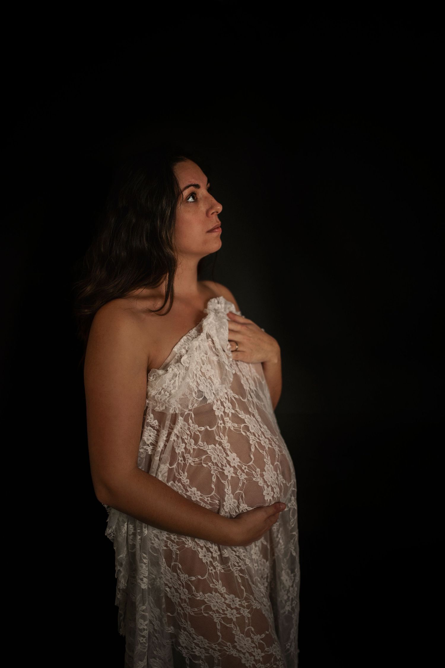Artistic maternity portrait featuring white lace against dramatic dark backdrop.