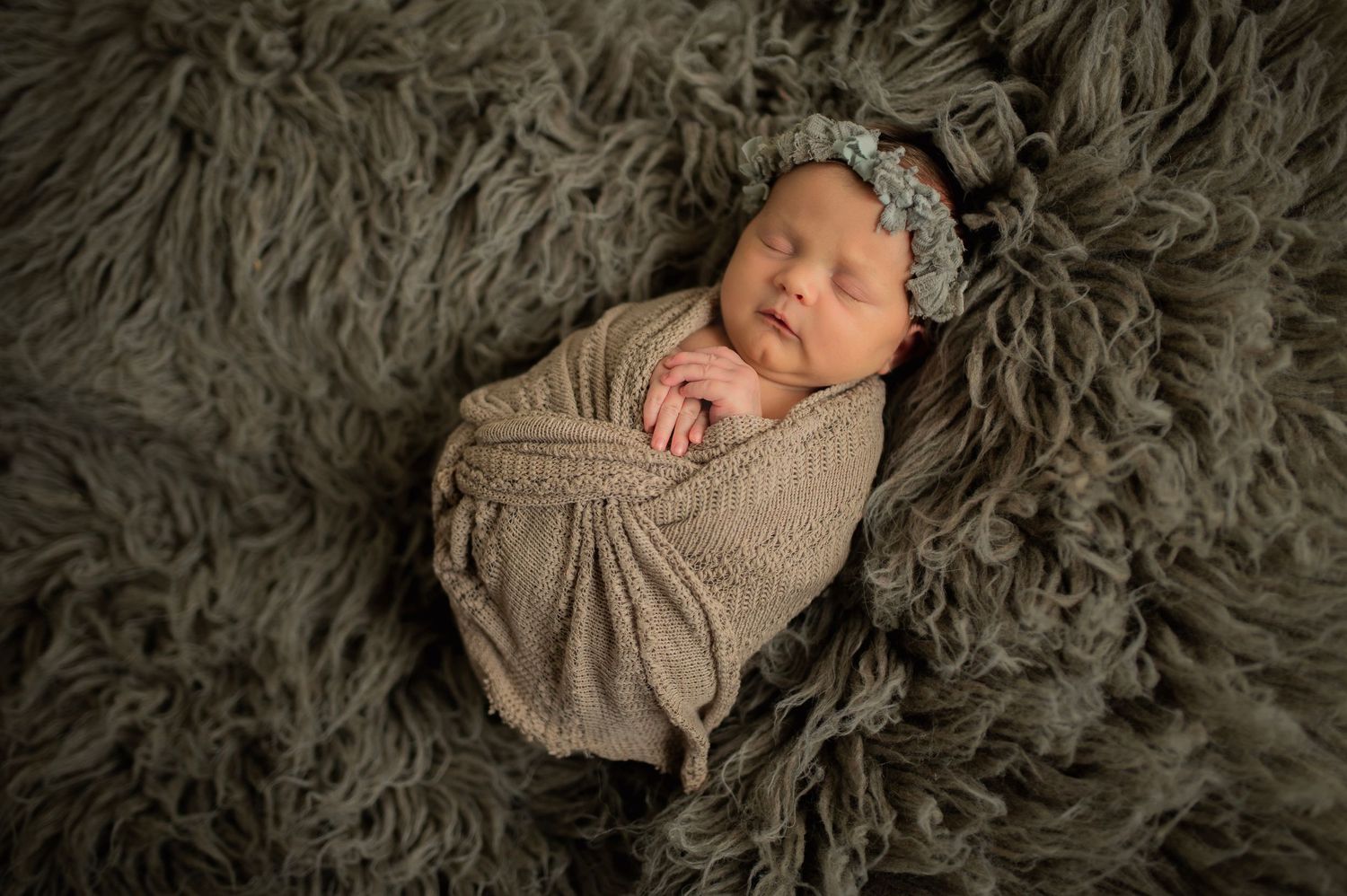 Sleeping newborn swaddled in beige wrap on textured gray blanket with lace headband.