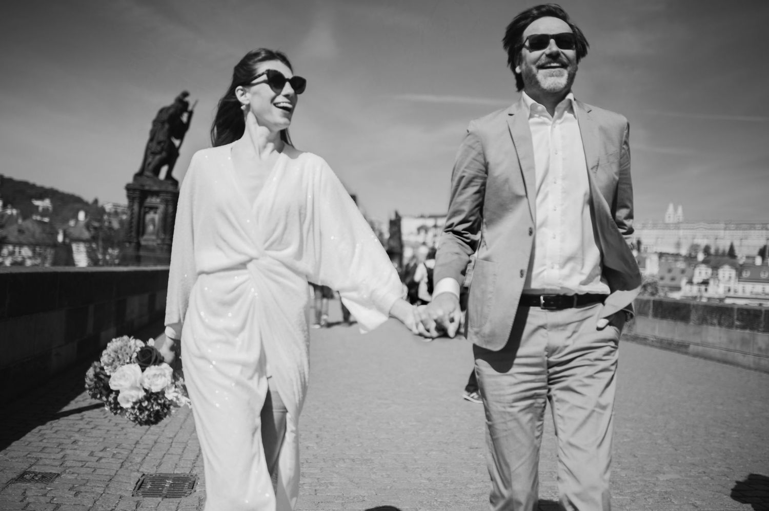 A black and white photo of a couple walking together on a bridge wearing sunglasses and formal attire.