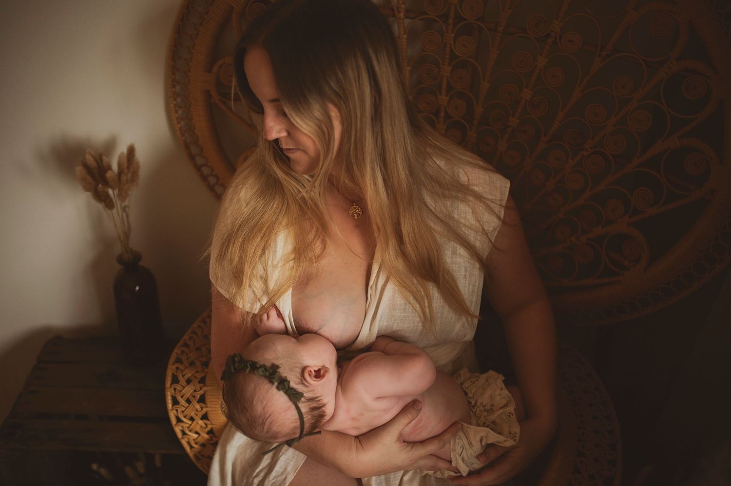 Intimate nursing portrait in warm tones with mother cradling infant in ornate peacock chair with ambient lighting.
