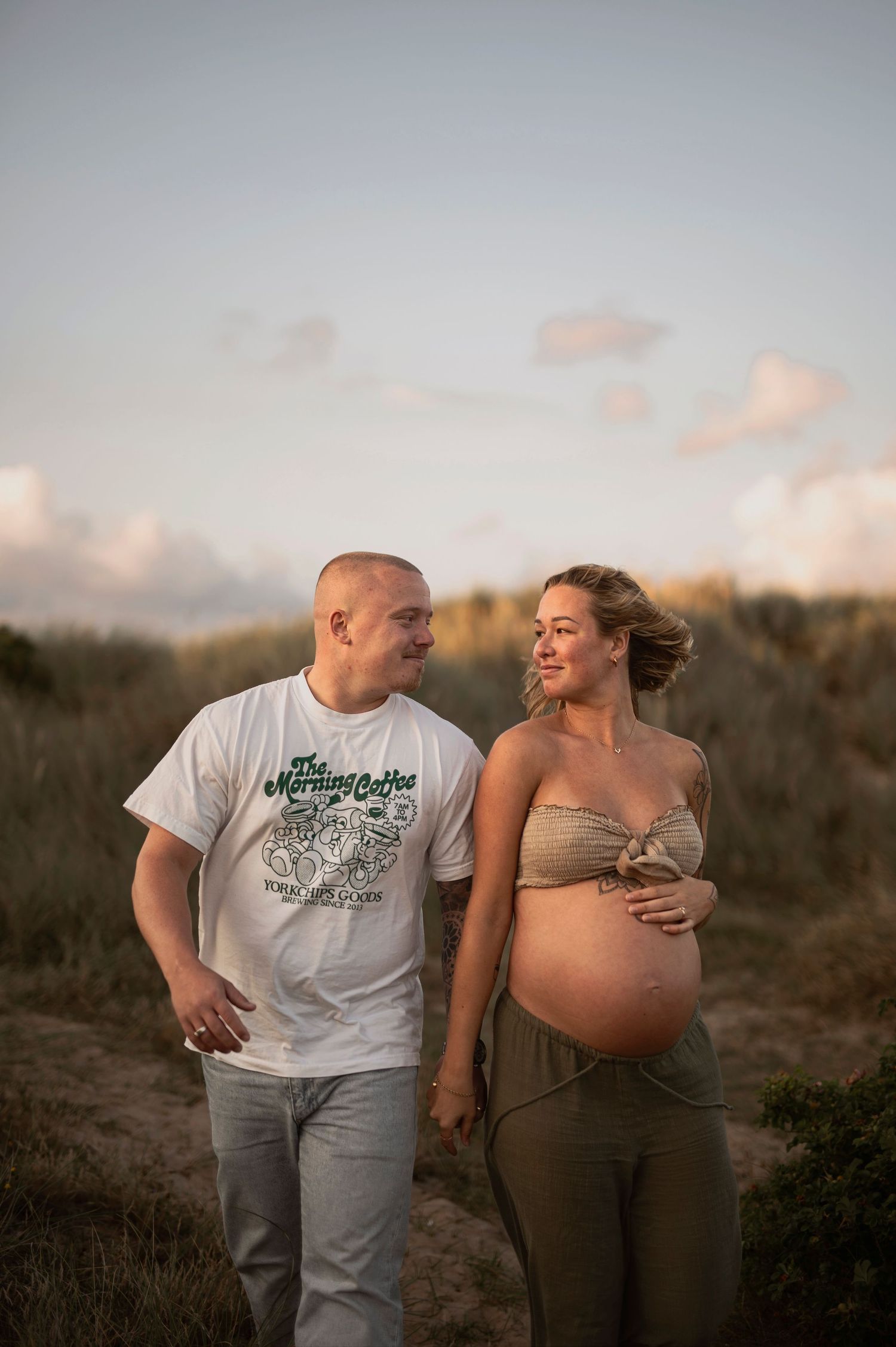 A pregnant couple shares an intimate moment outdoors at sunset while standing in a natural setting.