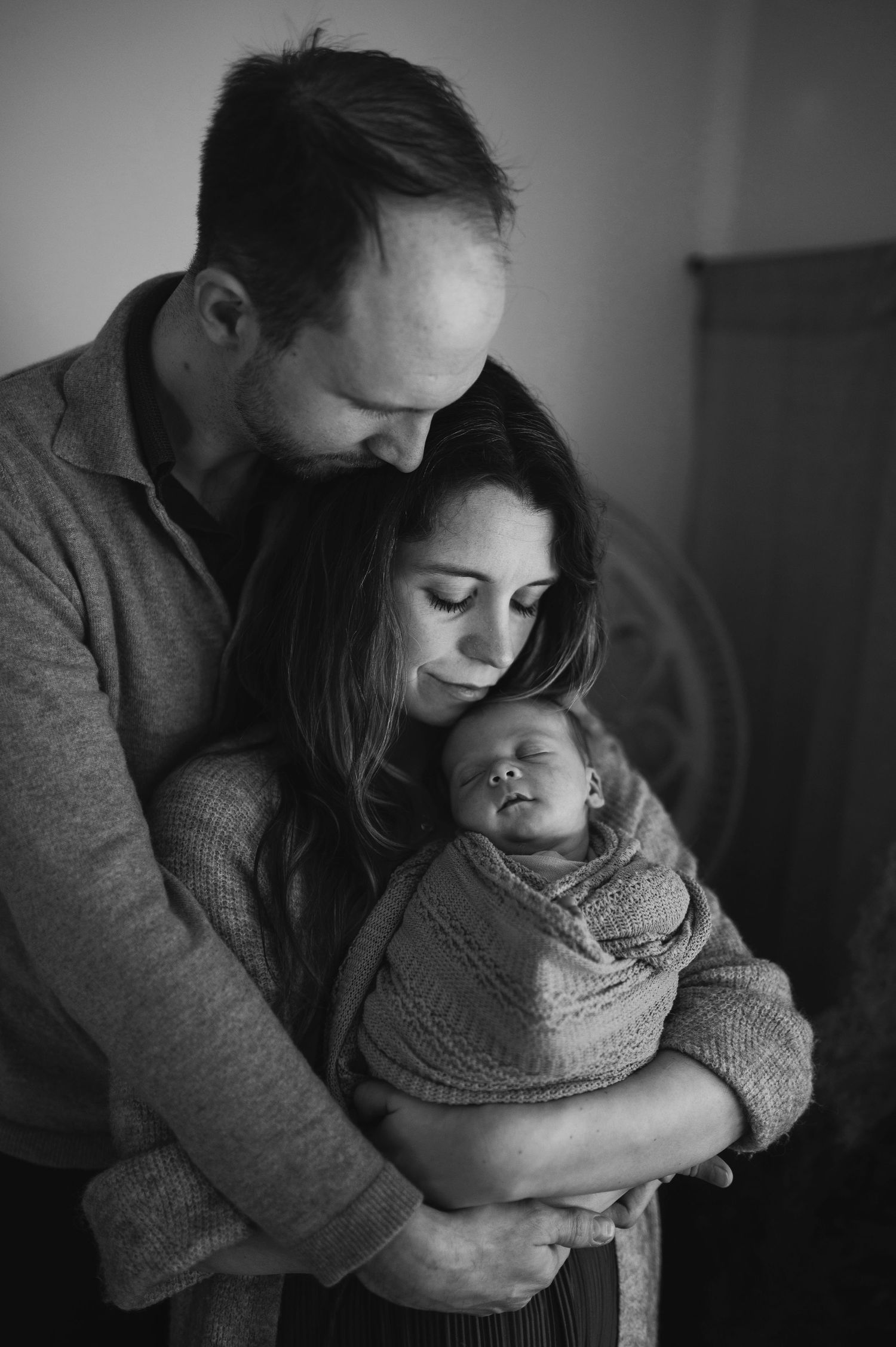 Atmospheric black and white image of parents holding their wrapped newborn in gentle embrace.