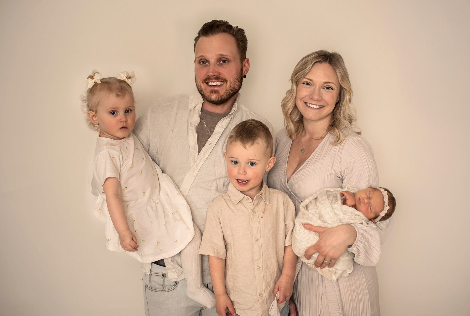 A family of five wearing neutral beige and white outfits poses together against a beige studio backdrop.