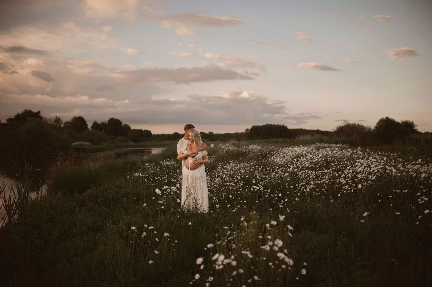 A couple embraces in a golden-hour meadow filled with white wildflowers under a dusky summer sky.