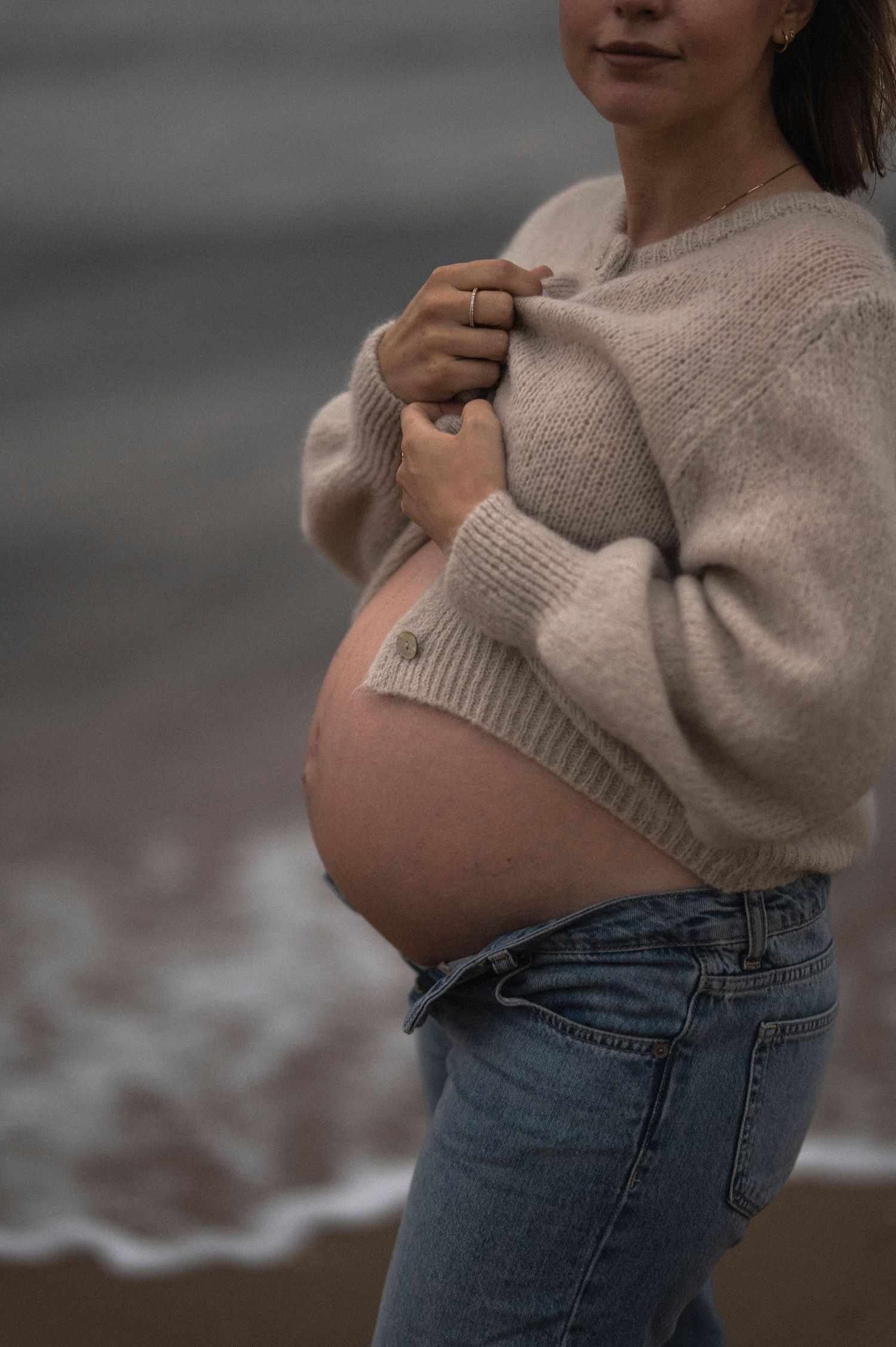 A side view of a pregnant woman in a beige sweater standing by the ocean.