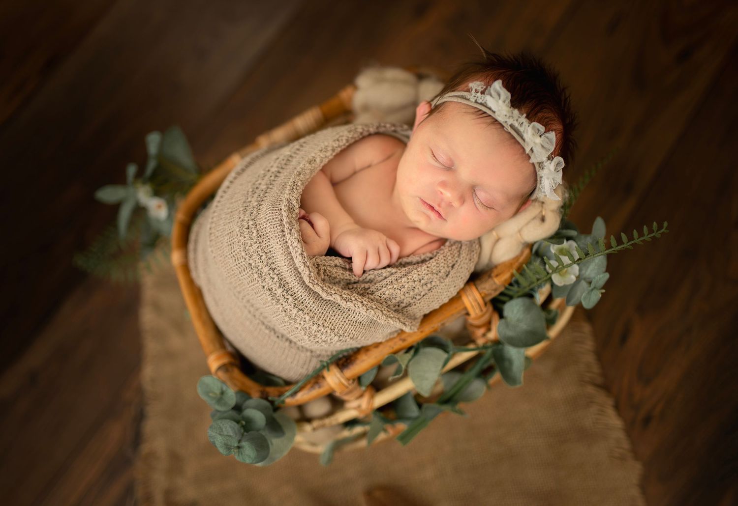 Sleeping newborn in basket surrounded by eucalyptus leaves and wearing knit wrap.