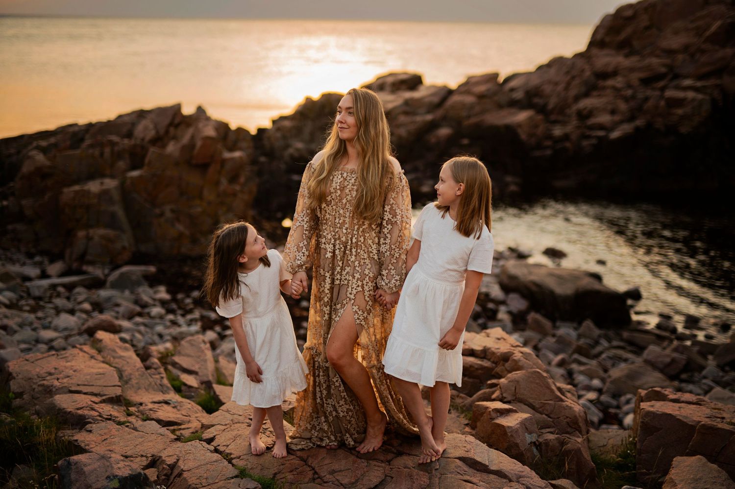 Family in white and gold dresses posing on rocky shore during golden sunset.