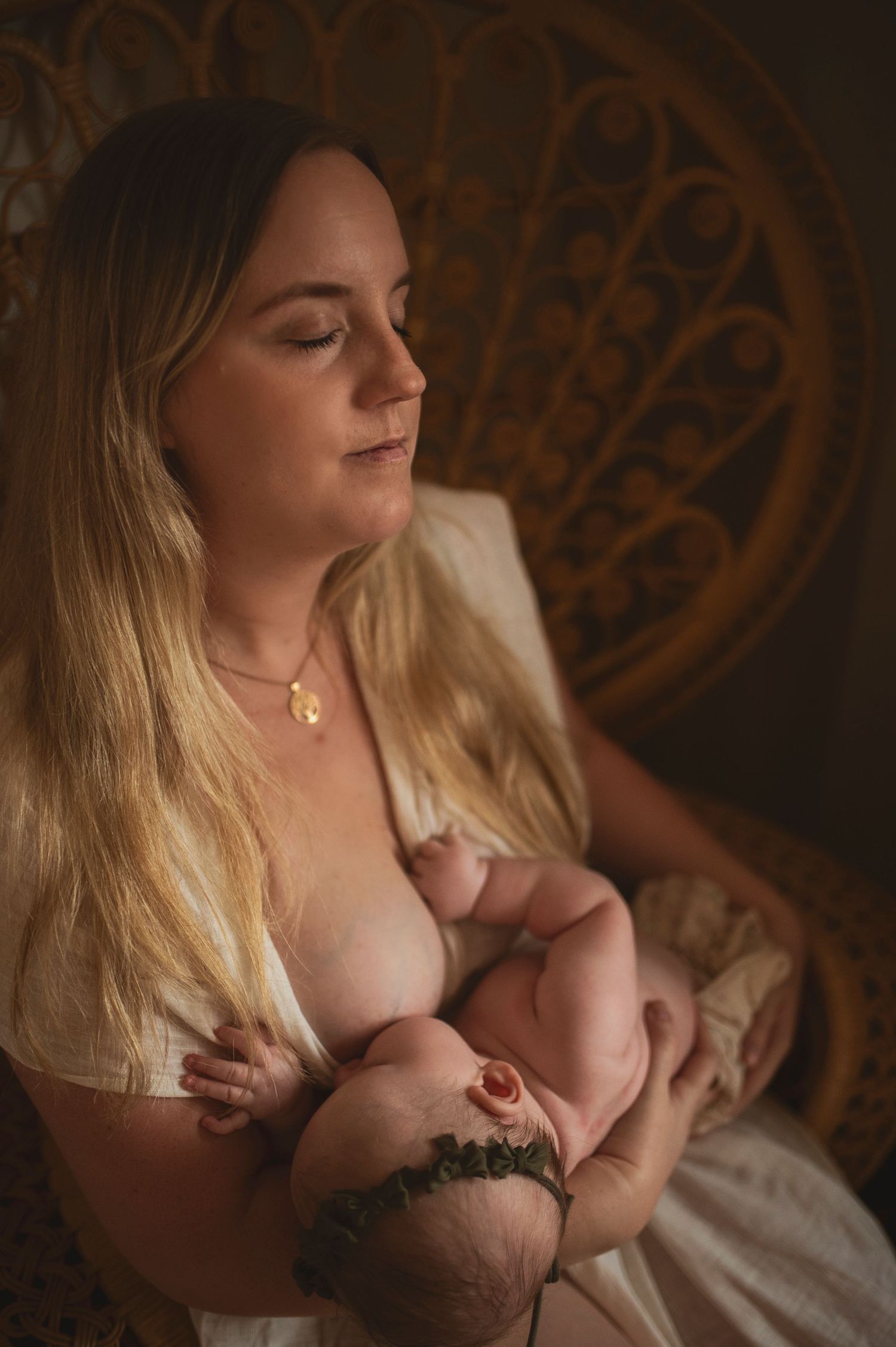 Artistic motherhood photography capturing intimate nursing moment in warm ambient light with woven chair backdrop.